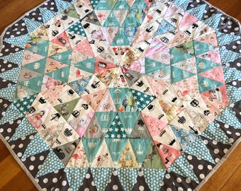 May include: A large, octagonal, quilted play mat with a gray and white polka dot border. The center of the mat features a colorful patchwork design with a variety of fabrics, including pink, blue, and green. The patchwork design is made up of triangles and stars.