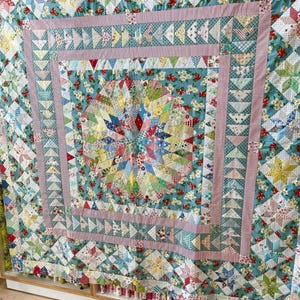 May include: A handmade quilt with a central starburst design in a variety of colorful fabrics. The quilt features a patchwork of floral, geometric, and striped patterns in shades of blue, red, pink, and yellow. The quilt is displayed on a wooden surface.