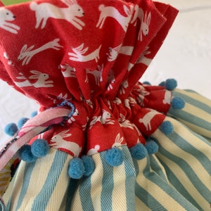 May include: A red fabric bag with a white bunny print, blue pom-poms, and a blue and white striped fabric base.