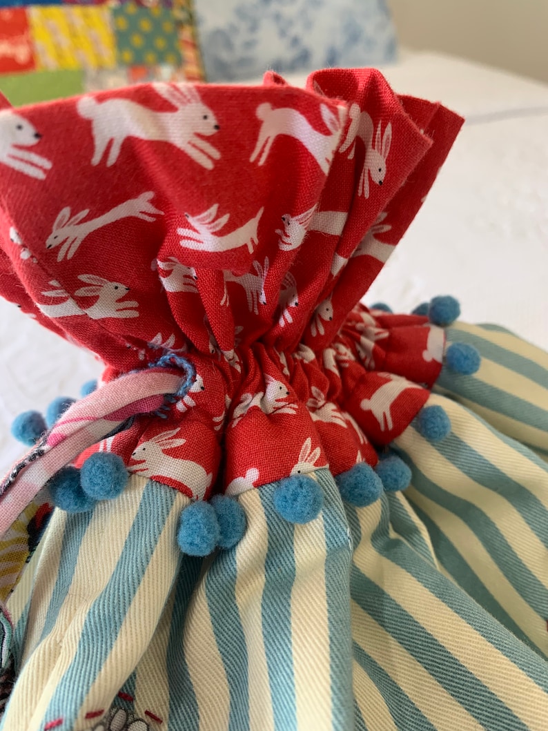 May include: A red fabric bag with a white bunny print, blue pom-poms, and a blue and white striped fabric base.