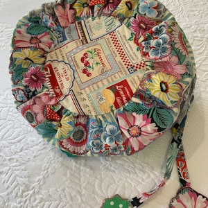 May include: A round, floral patterned cushion with a drawstring closure and a green and white polka dot tassel. The cushion is made of fabric with a vintage-inspired design featuring cherries, lemons, and flowers.