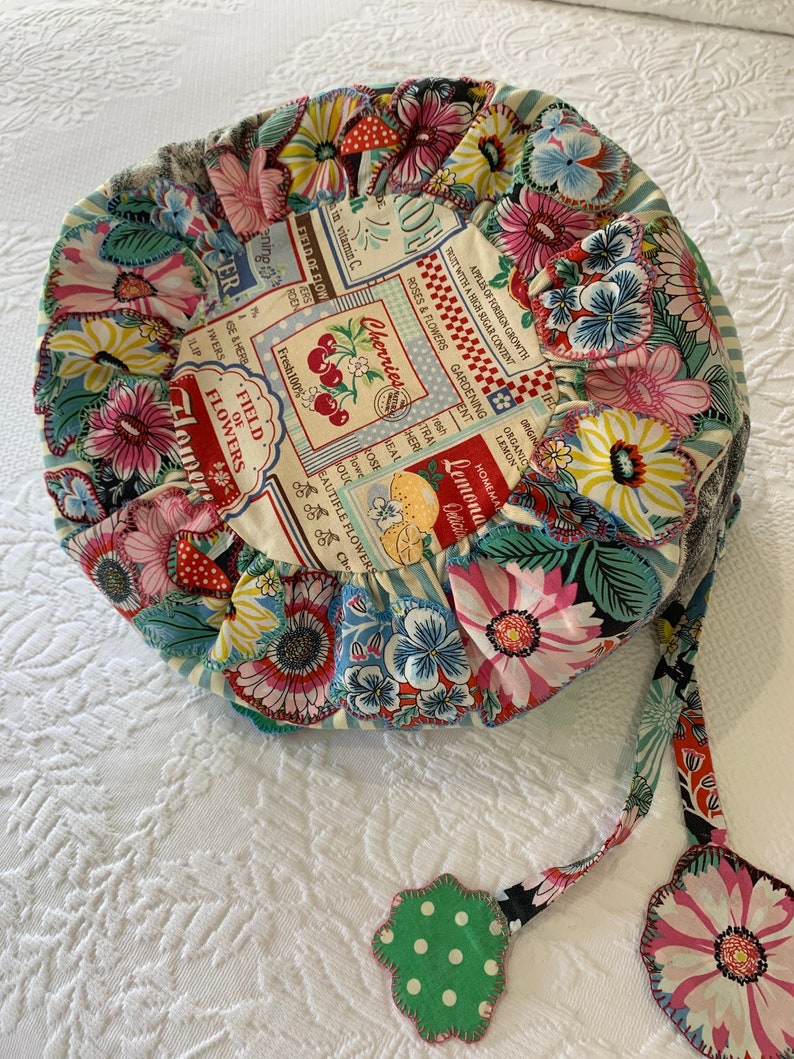 May include: A round, floral patterned cushion with a drawstring closure and a green and white polka dot tassel. The cushion is made of fabric with a vintage-inspired design featuring cherries, lemons, and flowers.
