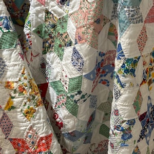 May include: Close-up of a vintage quilt with a star pattern. The quilt features white fabric with colorful star shapes made from various patterned fabrics in shades of red, green, blue, and pink. The quilt is folded, showing its texture and design.