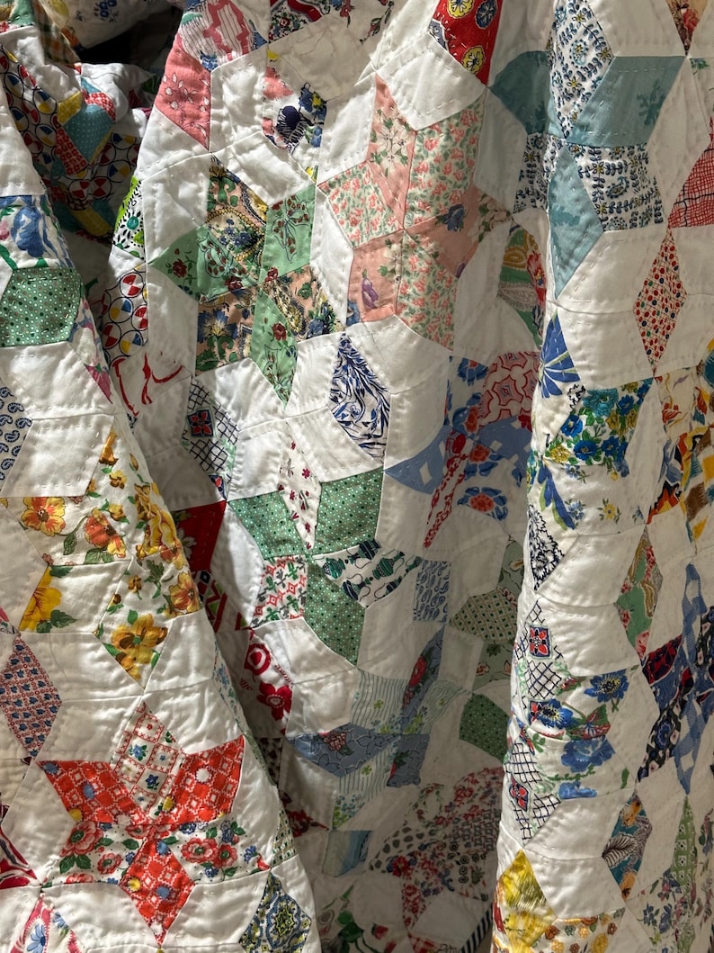 May include: Close-up of a vintage quilt with a star pattern. The quilt features white fabric with colorful star shapes made from various patterned fabrics in shades of red, green, blue, and pink. The quilt is folded, showing its texture and design.