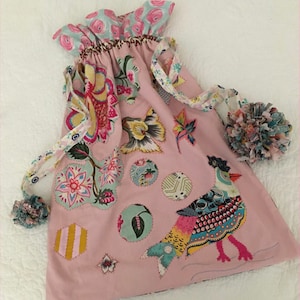 May include: A pink fabric bag with a drawstring closure. The bag is decorated with colourful floral and bird appliques. The bag has a ruffled top and is made of a soft, cotton fabric.