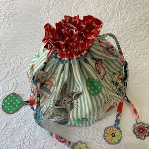 May include: A blue and white striped drawstring bag with a red and white bunny print. The bag has a ruffled top and is decorated with colorful fabric flowers.