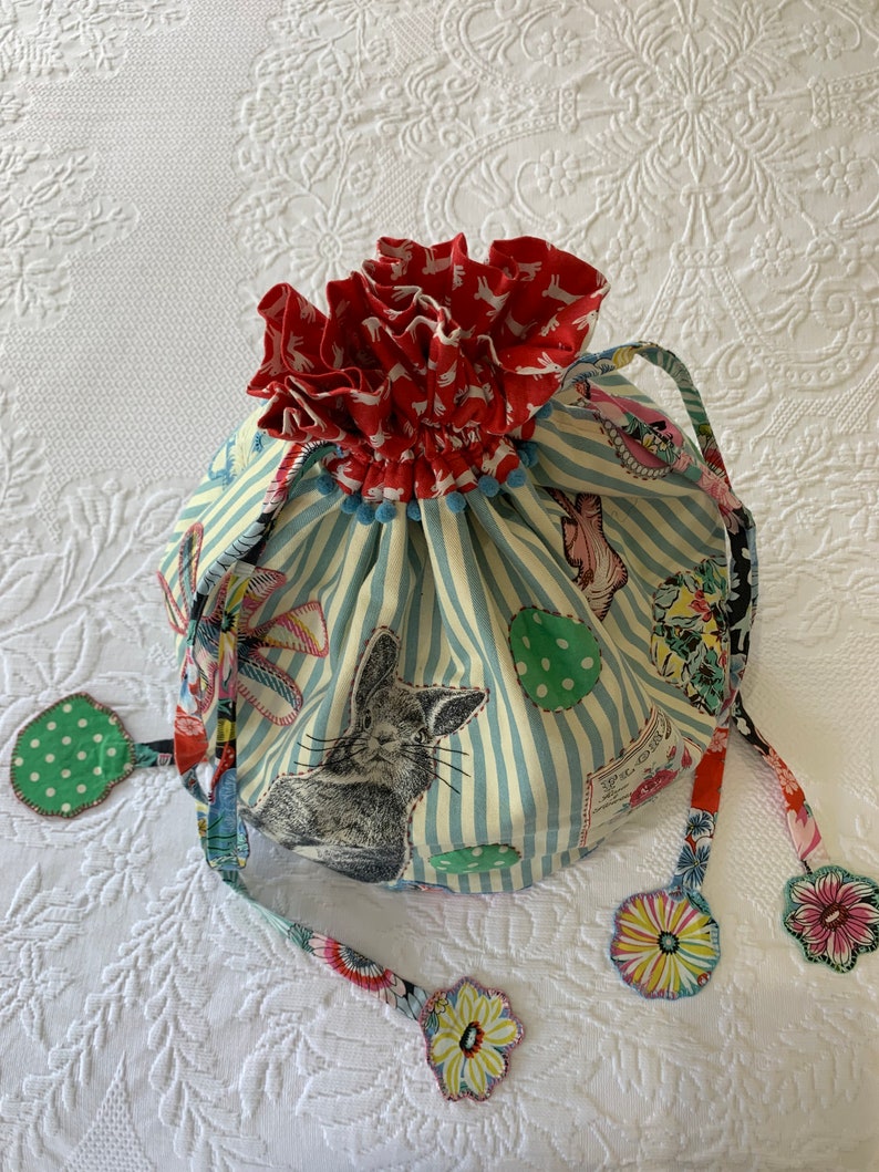 May include: A blue and white striped drawstring bag with a red and white bunny print. The bag has a ruffled top and is decorated with colorful fabric flowers.