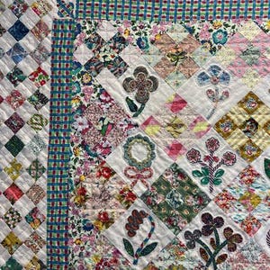 May include: A white quilt with a patchwork design featuring various floral patterns in shades of pink, green, blue, and yellow. The quilt is edged with a blue and green gingham border.