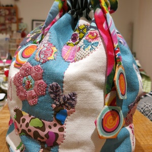 May include: A blue and white drawstring bag with a patchwork design featuring various colorful fabrics, including a pink floral pattern, a brown and white spotted pattern, and a red and yellow swirl pattern.