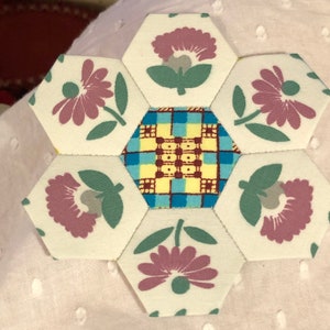 May include: A patchwork quilt block with a hexagonal shape. The block is made of six fabric squares with a floral pattern and one fabric square with a plaid pattern. The floral fabric is white with pink flowers and green leaves. The plaid fabric is yellow, blue, and brown.