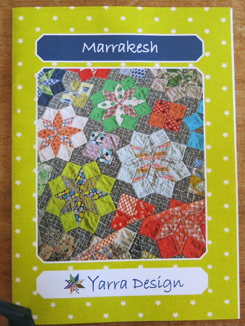 K&ouml;nnte beinhalten: Ein gr&uuml;nes, gelbes und rotes Quiltmuster mit dem Titel "Marrakesh" und dem Text "Yarra Design" auf gelbem Hintergrund mit wei&szlig;en Sternen.