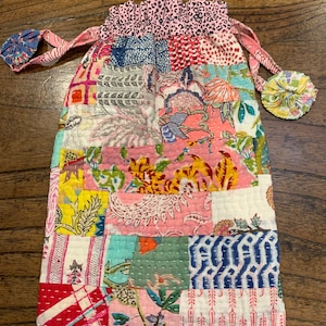 May include: A colourful patchwork drawstring bag with a pink, white and blue floral pattern. The bag has a gathered top and two fabric ties with decorative fabric balls.