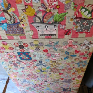 May include: A patchwork quilt with a floral design in the top section and a geometric pattern in the bottom section. The floral design features a bouquet of flowers in a pot with three figures in pink dresses. The geometric pattern is made up of various shapes and colors.