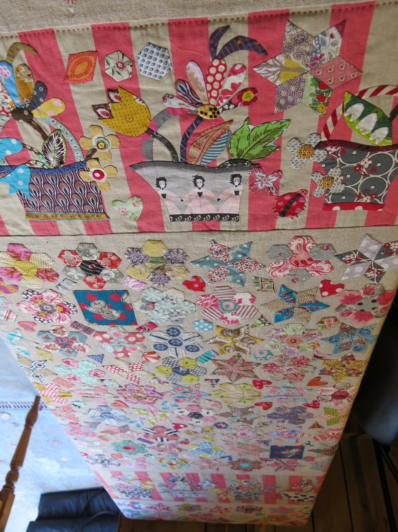 May include: A patchwork quilt with a floral design in the top section and a geometric pattern in the bottom section. The floral design features a bouquet of flowers in a pot with three figures in pink dresses. The geometric pattern is made up of various shapes and colors.