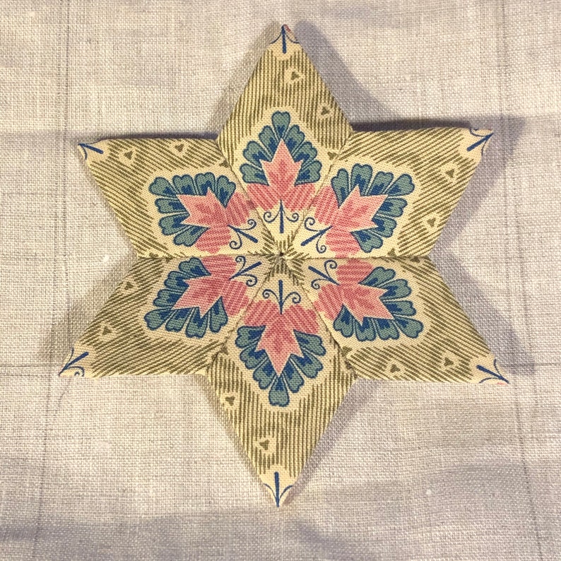 May include: A six-pointed star-shaped fabric patchwork with a repeating pattern of pink and blue leaves on a beige background.
