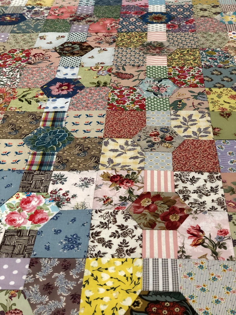 K&ouml;nnte beinhalten: Ein Patchwork-Deckenst&uuml;ck mit einer Vielzahl von floralen und geometrischen Mustern in Rosa-, Blau-, Gr&uuml;n- und Braunt&ouml;nen. Die Decke besteht aus vielen kleinen Quadraten und Sechsecken, die ein komplexes und interessantes Design ergeben.