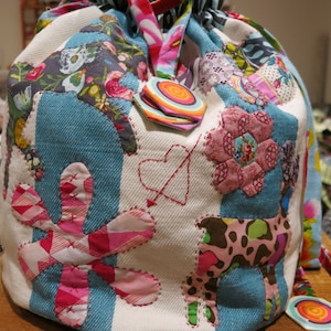 May include: A colorful patchwork drawstring bag with a variety of fabrics and patterns, including flowers, hearts, and geometric shapes. The bag is made of a white canvas with blue denim accents.