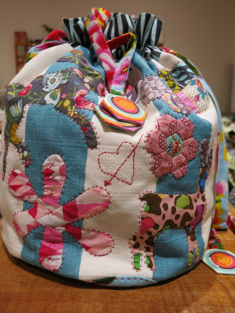 May include: A colorful patchwork drawstring bag with a variety of fabrics and patterns, including flowers, hearts, and geometric shapes. The bag is made of a white canvas with blue denim accents.