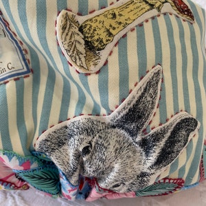 May include: A blue and white striped fabric bag with a variety of appliques, including a rabbit, a mushroom, and a floral design. The bag is made of a soft, textured fabric and has a unique, whimsical style.