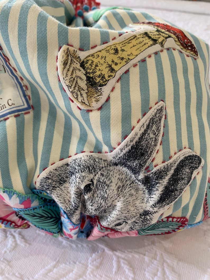 May include: A blue and white striped fabric bag with a variety of appliques, including a rabbit, a mushroom, and a floral design. The bag is made of a soft, textured fabric and has a unique, whimsical style.