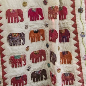 May include: A white fabric quilt with a red, white, and blue patchwork design. The quilt features a repeating pattern of elephants with different colored fabrics and designs. The quilt is edged with a red and white zig-zag pattern.