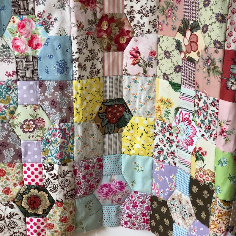 Puede incluir: Una colcha de retazos hecha de varias telas florales en tonos de rosa, azul, verde y amarillo. La colcha est&aacute; hecha de formas hexagonales y est&aacute; cosida con hilo blanco.