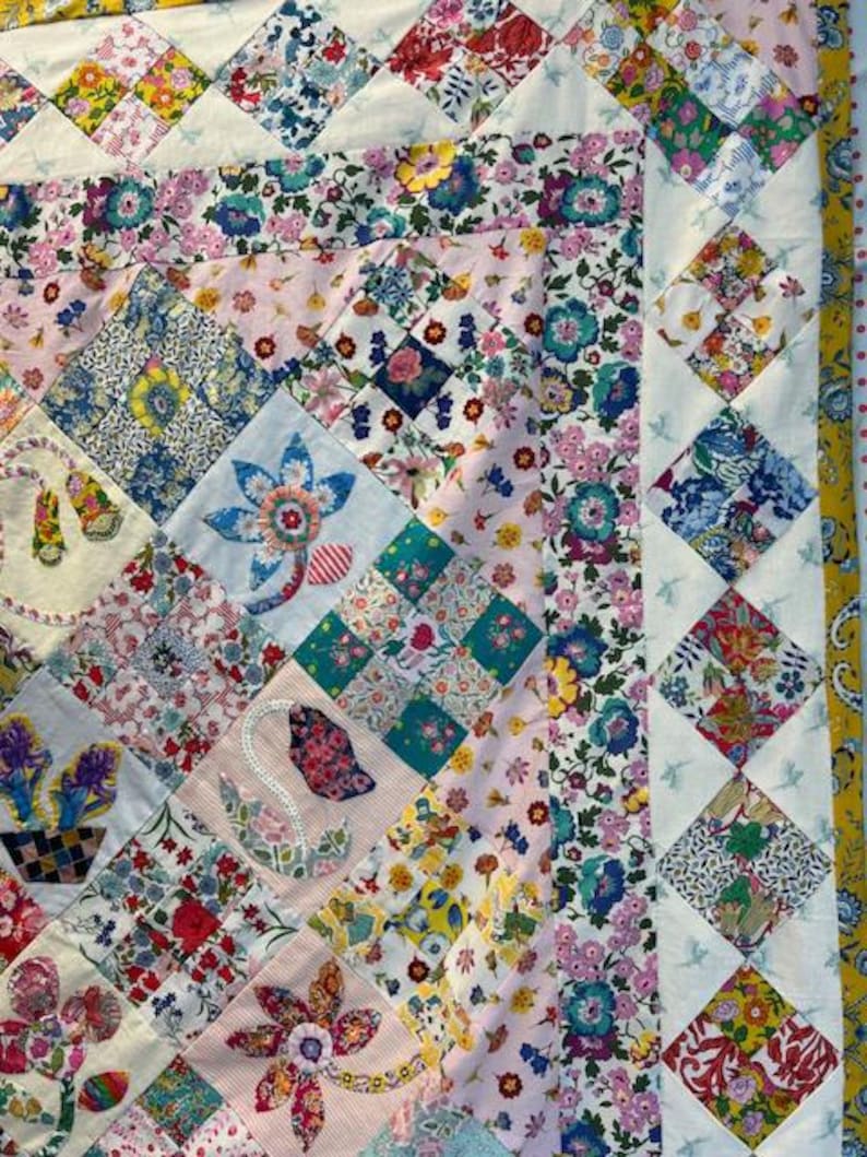 May include: A patchwork quilt with a floral pattern in shades of pink, blue, and yellow. The quilt is made up of squares of different fabrics, each with a unique design. The quilt is edged with a white border and a blue and white diamond pattern.