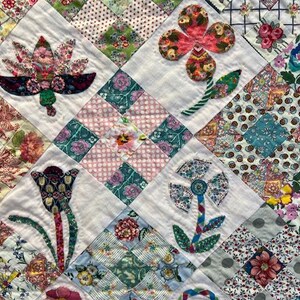 May include: A white quilt with a patchwork design featuring various floral motifs. The quilt is made with different fabrics in shades of pink, green, blue, and white. The flowers are appliqued and have a vintage style.