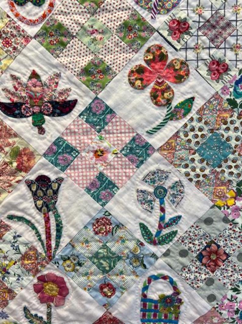 May include: A white quilt with a patchwork design featuring various floral motifs. The quilt is made with different fabrics in shades of pink, green, blue, and white. The flowers are appliqued and have a vintage style.
