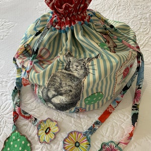 May include: A striped fabric drawstring bag with a red ruffled top and a cat print. The bag has four colorful fabric tassels attached to the drawstrings.