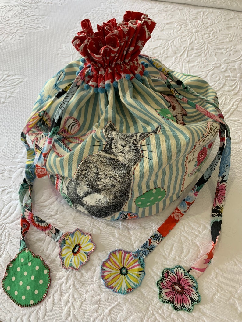 May include: A striped fabric drawstring bag with a red ruffled top and a cat print. The bag has four colorful fabric tassels attached to the drawstrings.
