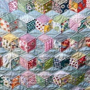 Puede incluir: Una colcha hecha de cuadrados de tela coloridos cosidos juntos en un patrón de cubos 3D. La colcha está hecha de una variedad de telas, incluyendo estampados florales, lunares, rayas y patrones geométricos. La colcha es de color azul claro con un patrón de cuadrícula blanca.