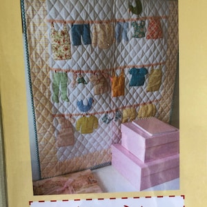 May include: A quilted wall hanging with a clothesline design featuring various colorful clothes and a bird. The wall hanging is displayed on a white wall with pink boxes stacked on a table below. The text "Trish Harper Designs" is displayed at the bottom of the image.