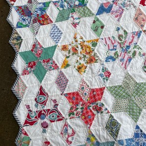 May include: Close-up of a handmade quilt featuring a star pattern. The quilt showcases a variety of colorful floral and geometric patterned fabrics, including red, green, blue, and yellow, set against a white background. The edges are finished with a striped border.