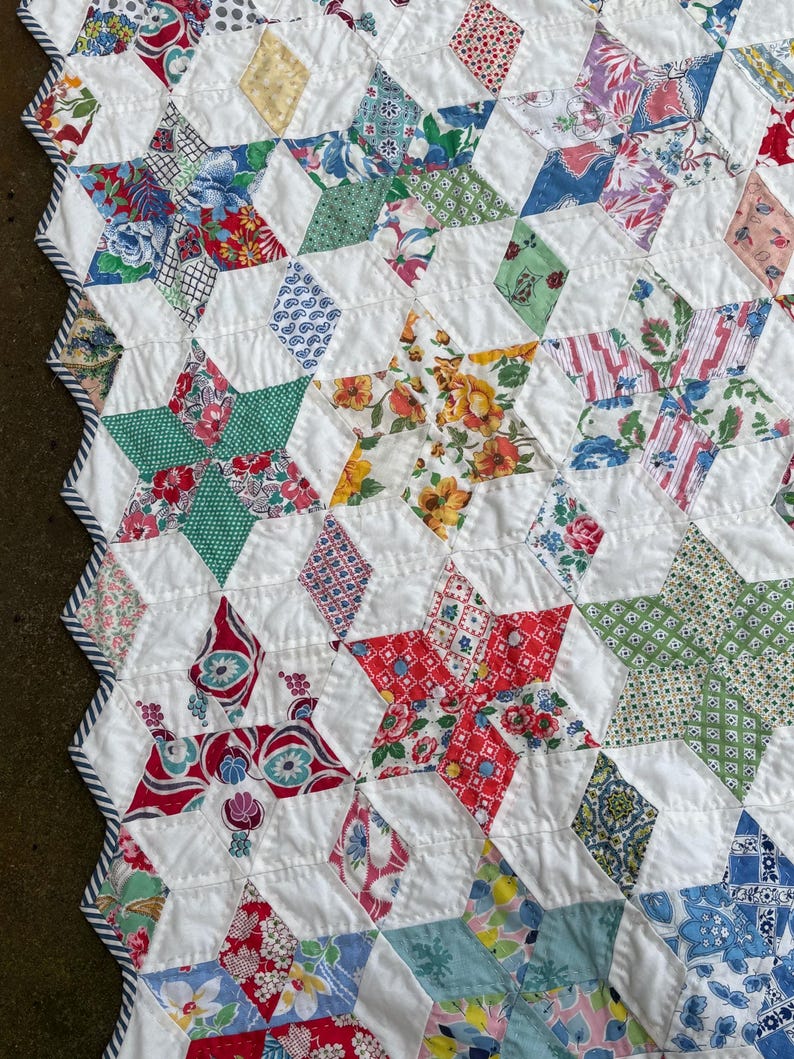 May include: Close-up of a handmade quilt featuring a star pattern. The quilt showcases a variety of colorful floral and geometric patterned fabrics, including red, green, blue, and yellow, set against a white background. The edges are finished with a striped border.