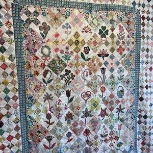 May include: A white quilt with a patchwork design featuring various floral patterns and colors. The quilt is bordered with a blue and white checkered fabric.
