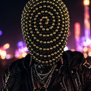 Op de afbeelding: Een zwart masker bedekt met goudkleurige bollen. Het masker wordt gedragen met een zwarte leren jas en meerdere zilveren kettingen. De achtergrond is wazig met kleurrijke lichten, wat een nachtelijke setting suggereert.