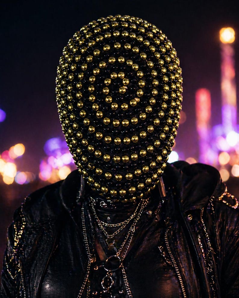 Puede incluir: Una m&aacute;scara negra cubierta de esferas doradas. La m&aacute;scara se usa con una chaqueta de cuero negra y m&uacute;ltiples cadenas plateadas. El fondo est&aacute; borroso con luces de colores, lo que sugiere un escenario nocturno.
