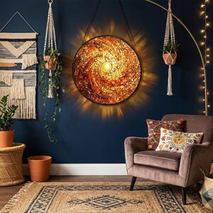 May include: A round, mosaic-style wall art piece with a spiral design in shades of orange and brown. The art is illuminated from behind, creating a warm, glowing effect. The piece is hanging on a dark blue wall, with a brown armchair and rug in the foreground.