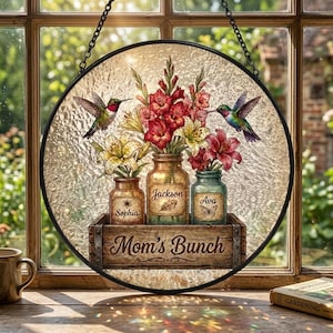 May include: Round stained glass window art featuring a floral arrangement in glass jars, with the names "Sophia", "Jackson", and "Ava". The words "Mom's Bunch" are on the wooden crate. Two hummingbirds are flying near the flowers.