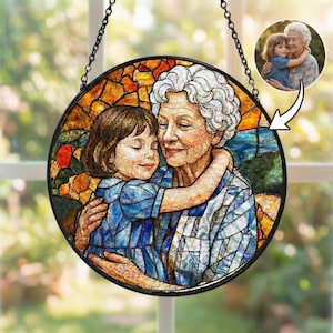 Könnte beinhalten: Rundes Buntglasfenster mit einer Großmutter und Enkelin, die sich umarmen. Das Kunstwerk hat einen schwarzen Metallrahmen und eine Kette zum Aufhängen. Das Bild ist ein farbenfrohes Mosaik aus Orange-, Gelb-, Blau- und Grüntönen mit einem kleineren Foto.