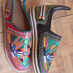 May include: A pair of colorful, embroidered leather slippers. One slipper is red with a green trim, the other is black. Both feature intricate geometric patterns in various colors, including yellow, blue, and pink.