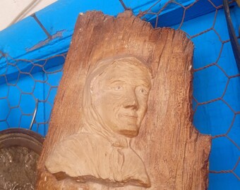 May include: A carved wooden relief sculpture of a person in a hooded garment. The sculpture is mounted on a piece of rough-hewn wood. The background includes a blue surface and a metal wire mesh.