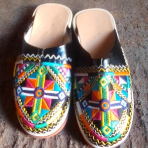 May include: A pair of black leather slippers with colorful geometric embroidery. The slippers feature a vibrant design with orange, blue, green, and pink accents. The soles are white and the interior is a light tan color.