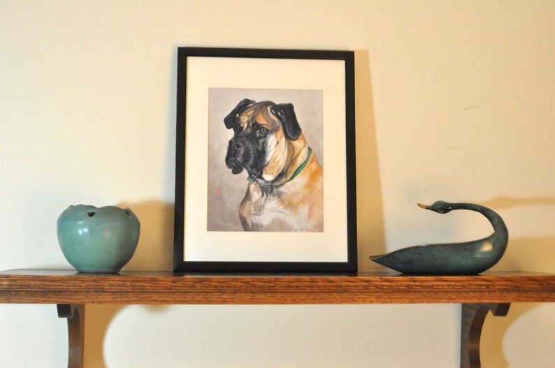 May include: A framed painting of a brown and black dog with a green collar. The painting is on a wooden shelf with a teal ceramic bowl and a bronze swan figurine.