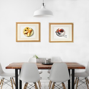 May include: Two framed watercolor paintings, one depicting a plate of pastries and the other a cup of tea with a saucer. The paintings are hanging on a white wall above a wooden table with four white chairs.