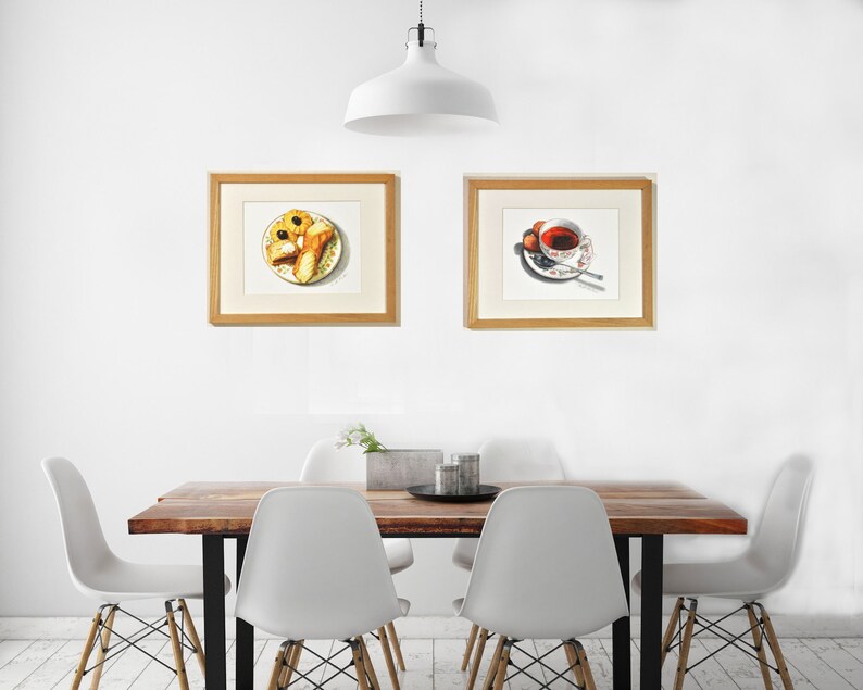 May include: Two framed watercolor paintings, one depicting a plate of pastries and the other a cup of tea with a saucer. The paintings are hanging on a white wall above a wooden table with four white chairs.