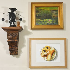 May include: A framed painting of a pond with lily pads and pink flowers, a framed drawing of pastries on a plate, and a wooden shelf with a carved figure holding a balancing pole with two baskets.