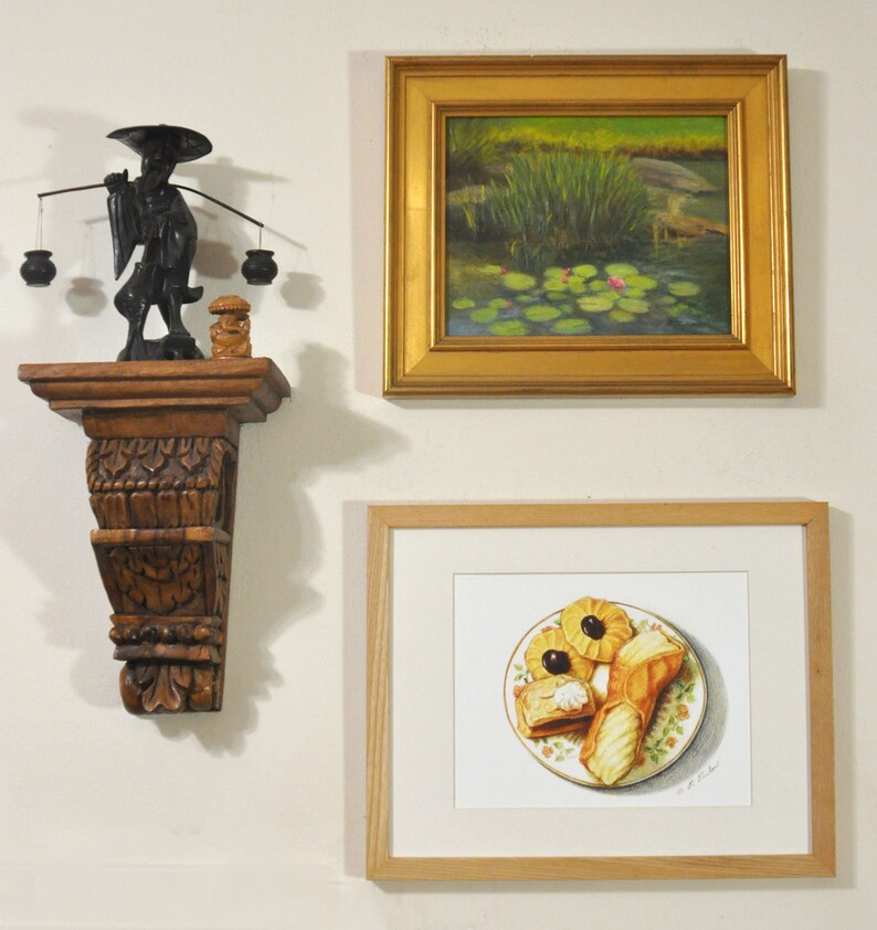 May include: A framed painting of a pond with lily pads and pink flowers, a framed drawing of pastries on a plate, and a wooden shelf with a carved figure holding a balancing pole with two baskets.