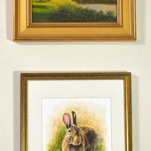 May include: A framed watercolor painting of a brown rabbit sitting in a field of green grass. The rabbit has pink ears and is looking directly at the viewer.